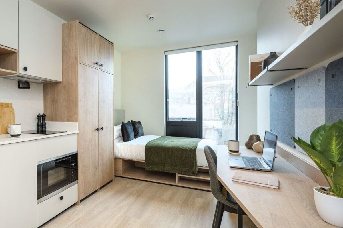 Une pièce de taille modeste comprenant un lit, un bureau avec un ordinateur portable, un petit coin cuisine et une grande fenêtre.