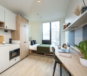 A compact room with a bed, a desk with a laptop, a small kitchen area, and a large window.