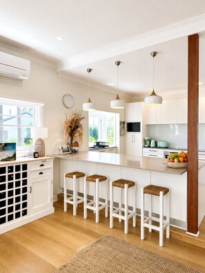 A modern kitchen with white cabinetry, a central island with stools, and a large window providing natural light. - Beautiful Hamptons style home, just a short walk from the Beach, shops & cafés. (Freshwater)