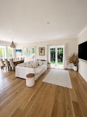A spacious living room with a large sofa, dining area, and a view of greenery through the windows. - Beautiful Hamptons style home, just a short walk from the Beach, shops & cafés. (Freshwater)