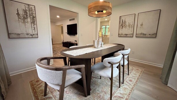 Uma sala de jantar com mesa de madeira, quatro cadeiras, luminária pendente e obras de arte nas paredes.