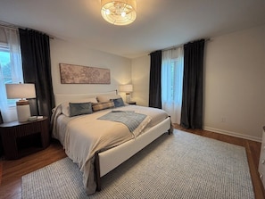A bedroom with a large bed, a bedside table, a lamp, and a window with curtains. - Covenanter Classic | Luxe East Side Retreat (Bloomington)
