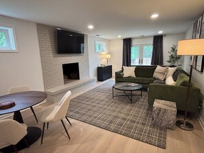 A modern living room with a fireplace, a flat-screen TV, a sofa, a coffee table, and a dining area with chairs. - Covenanter Classic | Luxe East Side Retreat (Bloomington)
