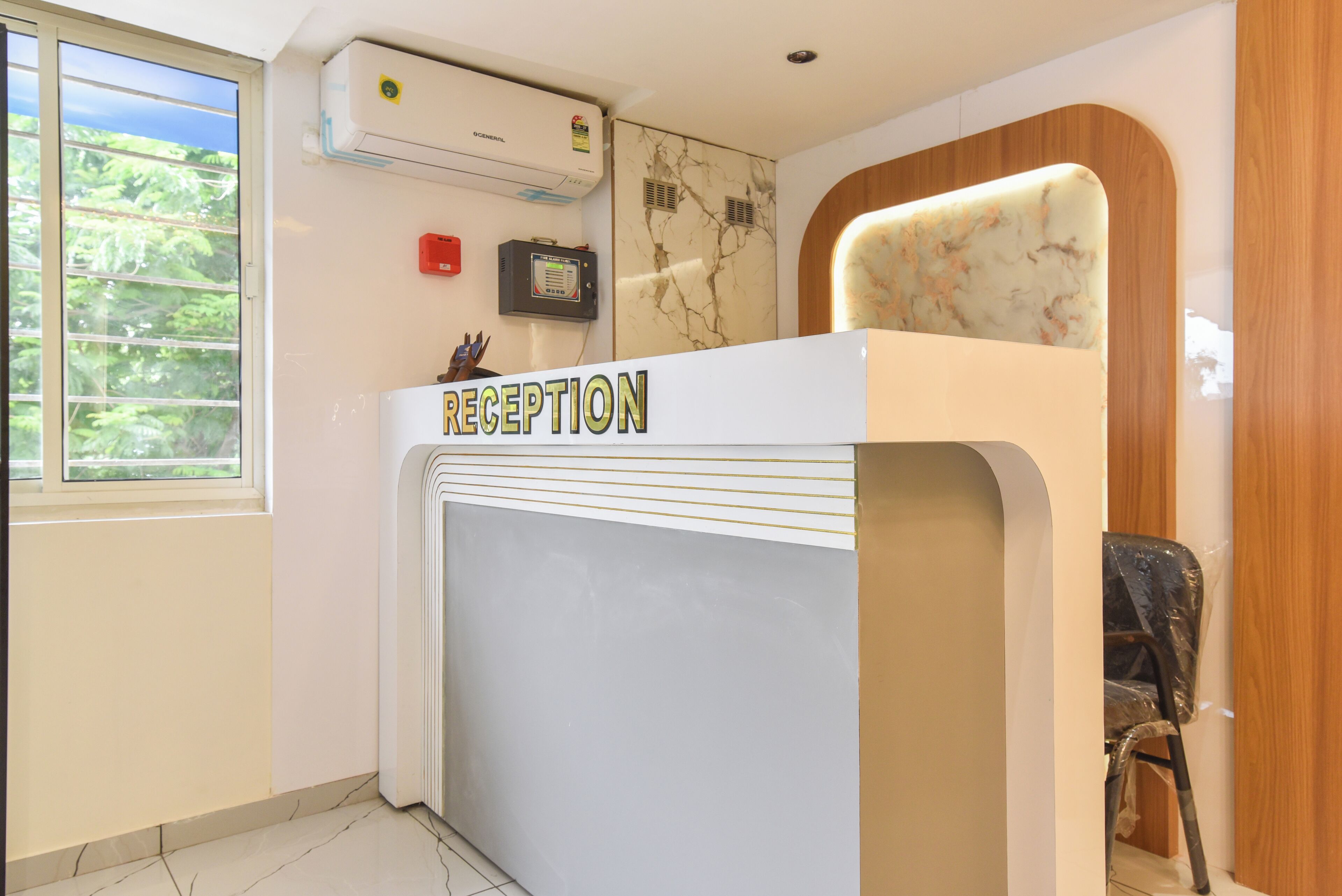 A reception desk with a 'RECEPTION' sign, an air conditioning unit above it, a wall-mounted fire alarm panel, and a window with a view of greenery.