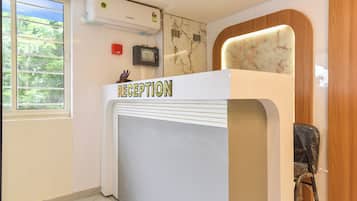 A reception desk with a 'RECEPTION' sign, an air conditioning unit above it, a wall-mounted fire alarm panel, and a window with a view of greenery.