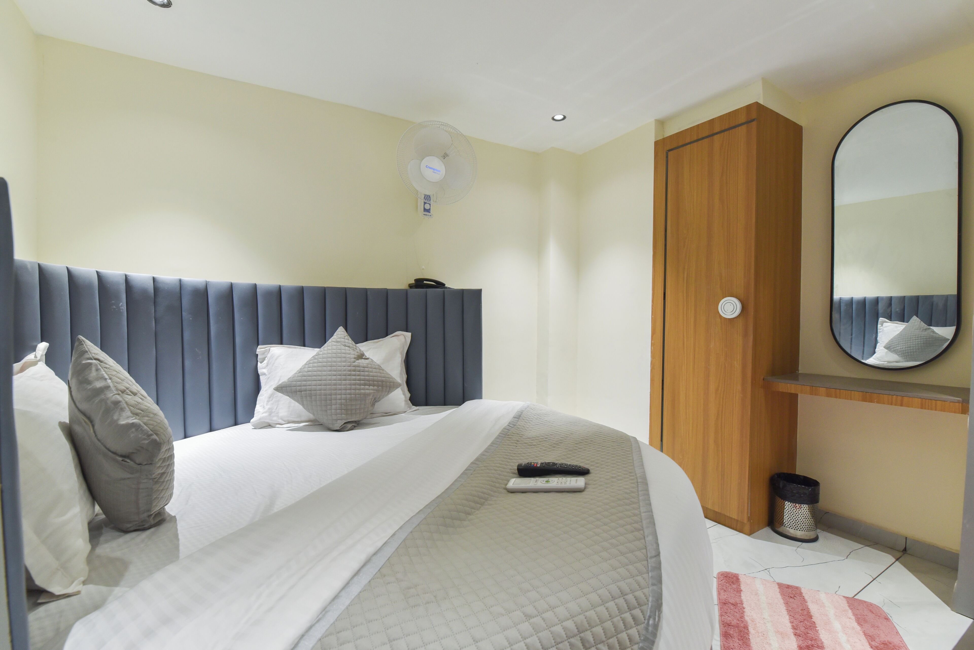Un dormitorio con cama, almohadas, un control remoto, un ventilador de pared, una puerta de madera y un espejo.