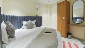 Un dormitorio con cama, almohadas, un control remoto, un ventilador de pared, una puerta de madera y un espejo.