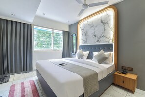 A modern bedroom with a large bed, a ceiling fan, a marble-patterned headboard, a bedside table with a phone, and a view of greenery through the window. - Hotel Prime Icon (Ahmedabad)