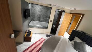 A modern bedroom with a large bed, a TV, a bathroom with a bathtub, and a wooden door.
