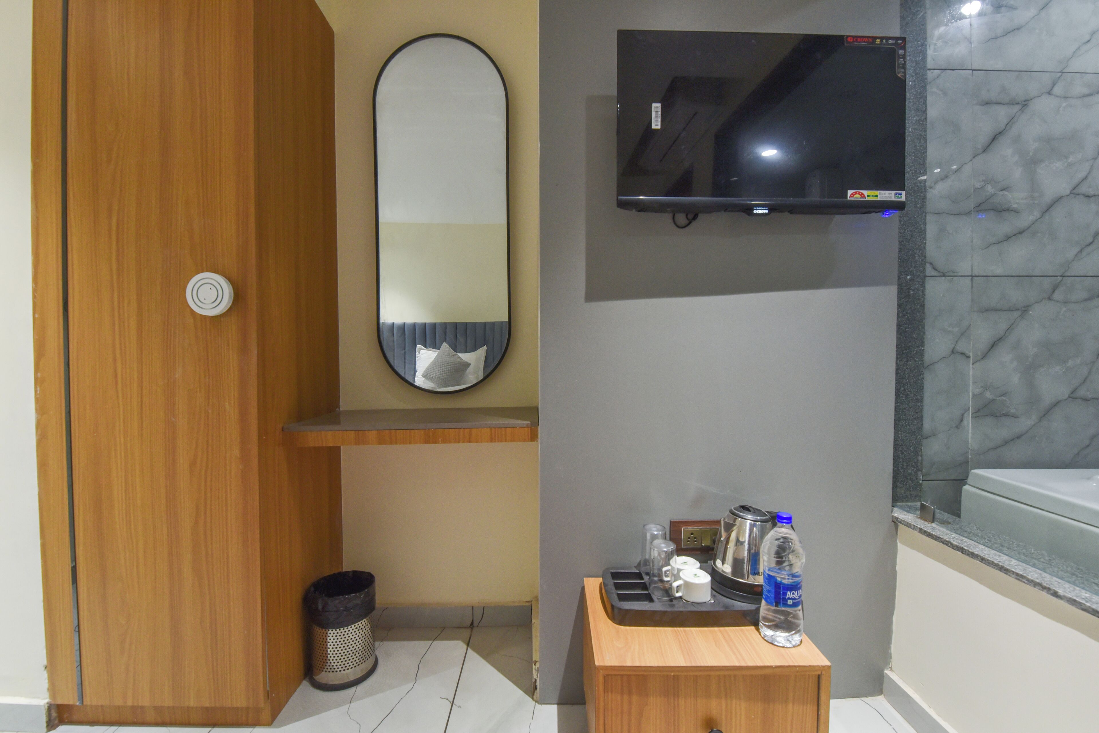 Un baño con puerta de madera, un espejo, un televisor montado, un tocador pequeño de madera con una botella de agua y un cesto de basura.