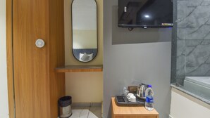Un baño con puerta de madera, un espejo, un televisor montado, un tocador pequeño de madera con una botella de agua y un cesto de basura.