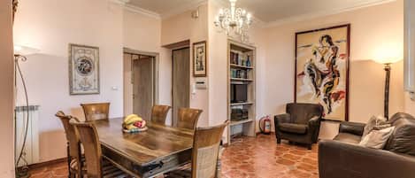 A dining area with a wooden table and chairs, a chandelier, and a bookshelf with books and decorative items.
