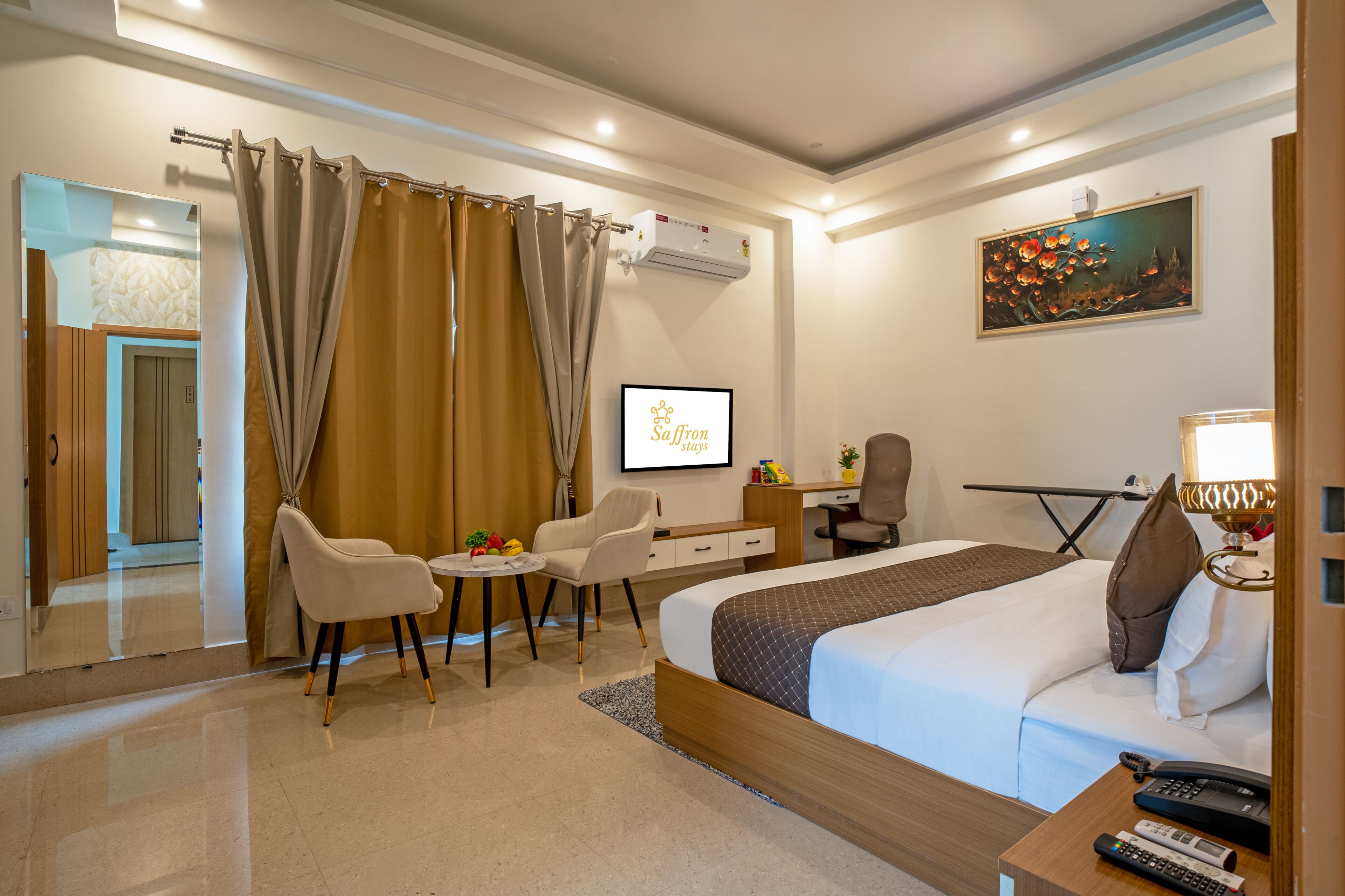 A hotel room with a bed, desk, chairs, a television, and a painting on the wall.