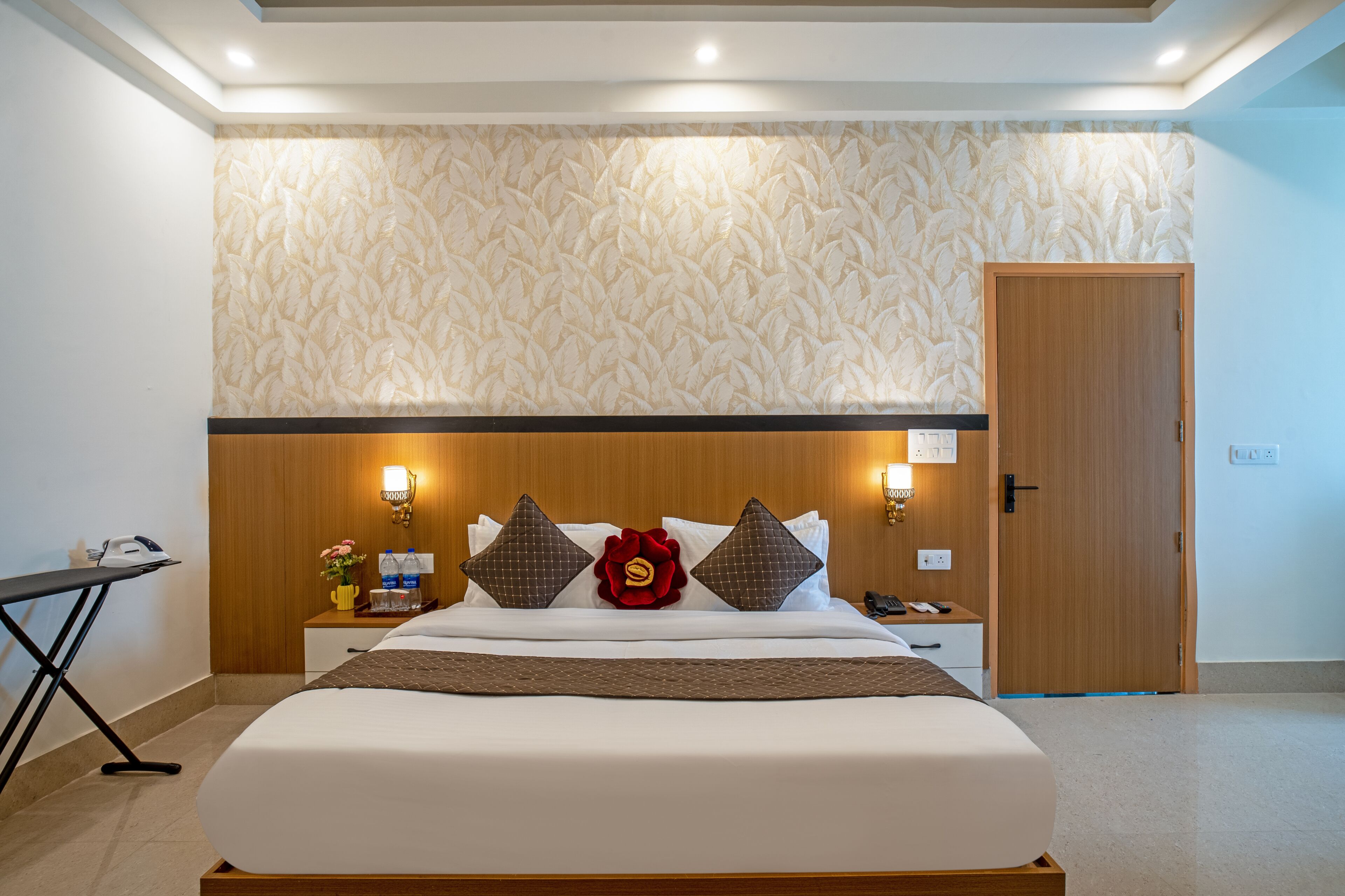 Een hotelkamer met een groot bed, een houten hoofdeinde, een nachtkastje met een lamp en een deur.