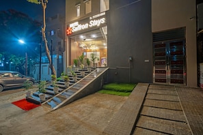 Entrada de hotel com escadas que levam à porta, plantas em vasos e um tapete vermelho.