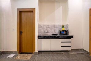 A compact kitchen with a wooden door, white cabinets, a marble backsplash, and a microwave on the countertop. - Hotel Saffron Stays (Noida)