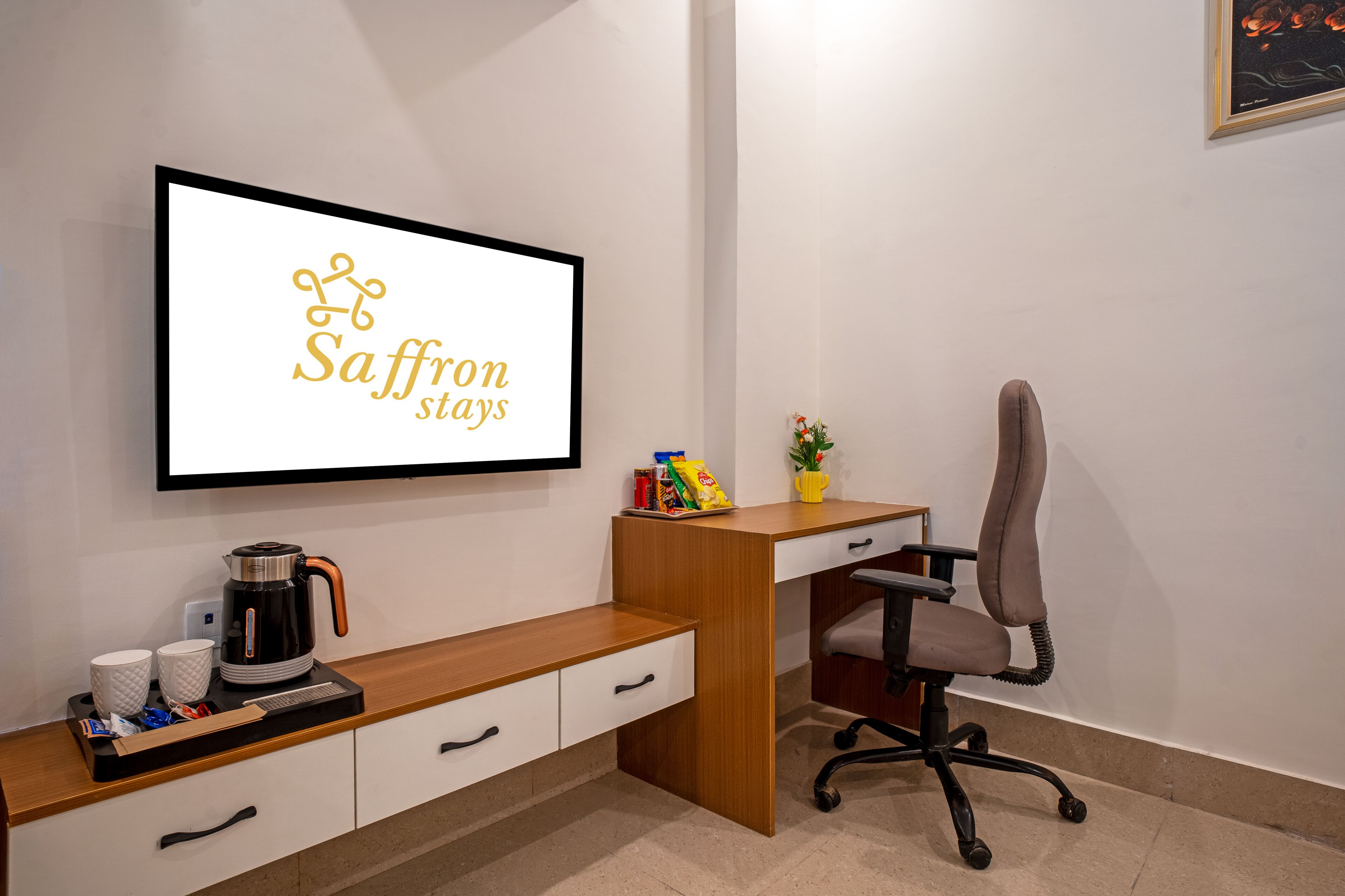 A hotel room with a desk, a chair, a coffee maker, and a framed sign that reads 'Saffron stays'.