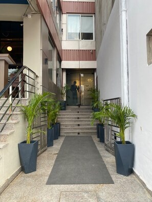 Entrada de un edificio con escaleras, plantas en macetas y una puerta con señalización.