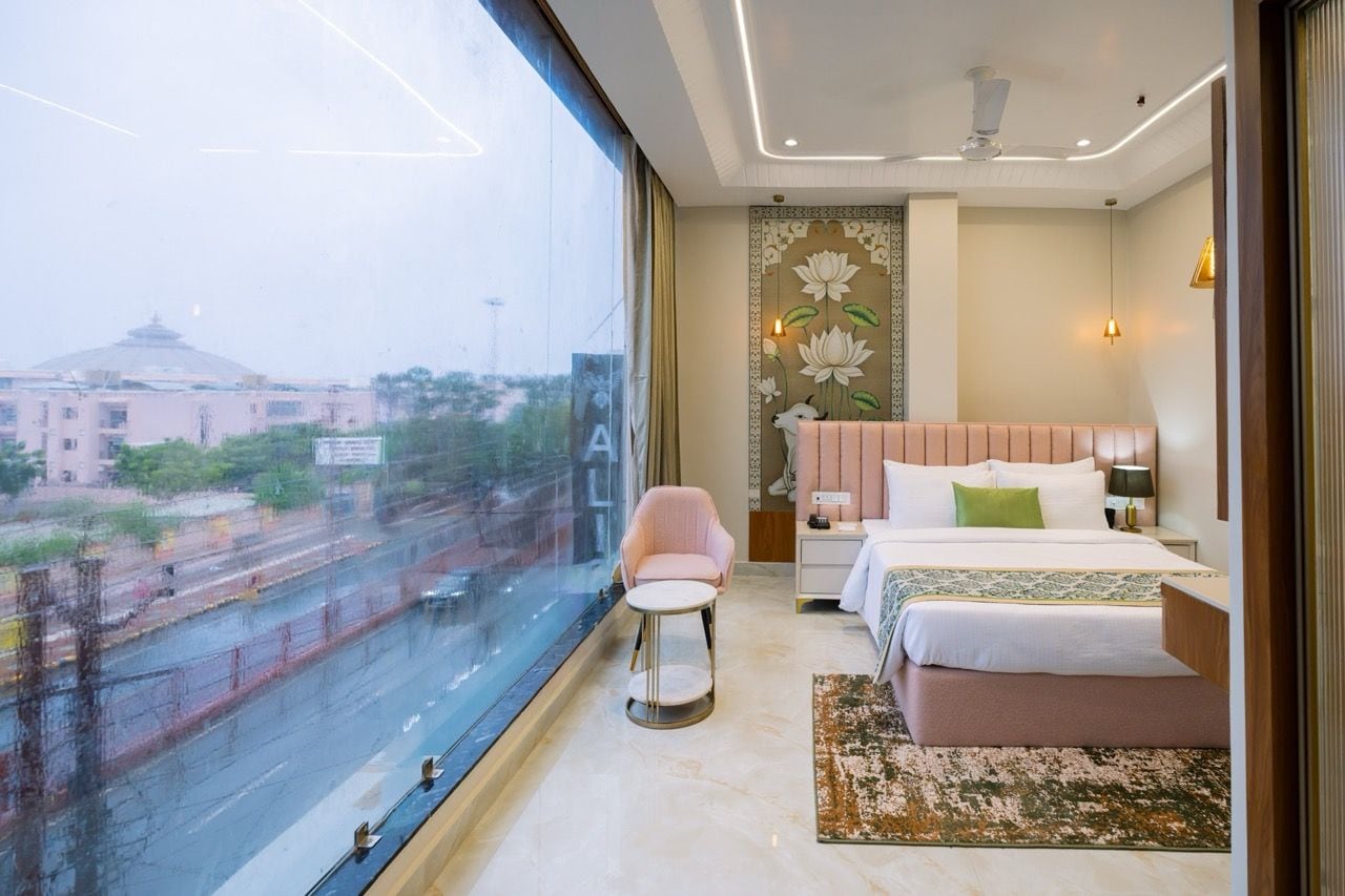 A hotel room with a large window, a bed, a bedside table, a chair, and a decorative wall panel.