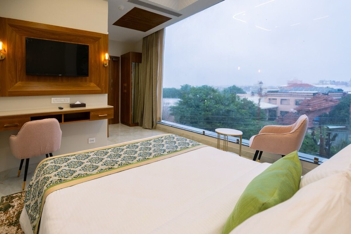 A hotel room with a large bed, a desk with a flat-screen TV, two chairs, and a view of the city through a large window.