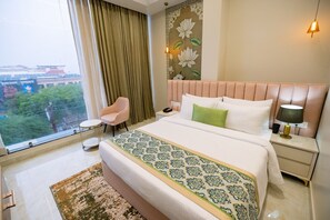 A hotel room with a large bed, a chair, a bedside table with a lamp, and a view of a cityscape through a window. - Aliste Hotel Prem Mandir Vrindavan (Vrindavan)