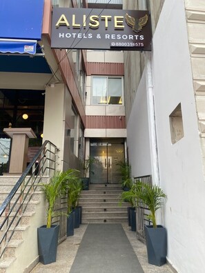A hotel entrance with a sign reading 'ALISTE HOTELS & RESORTS' and a phone number. There are steps leading up to the entrance, and potted plants are placed along the path.