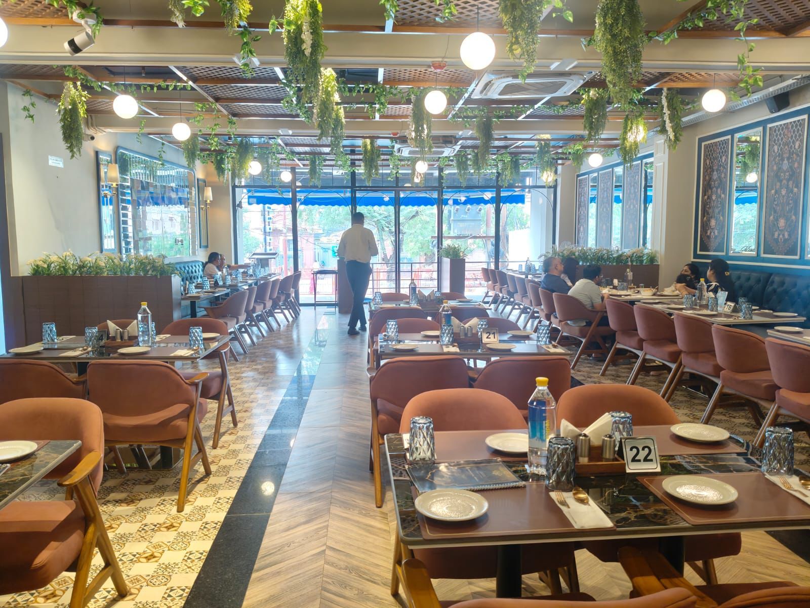 Un restaurante espacioso con varias mesas y sillas, piso con diseño y plantas colgantes.