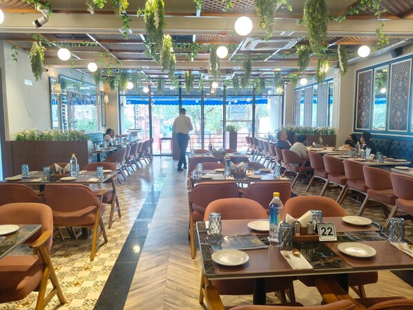 A spacious restaurant with multiple dining tables and chairs, a patterned floor, and hanging plants.