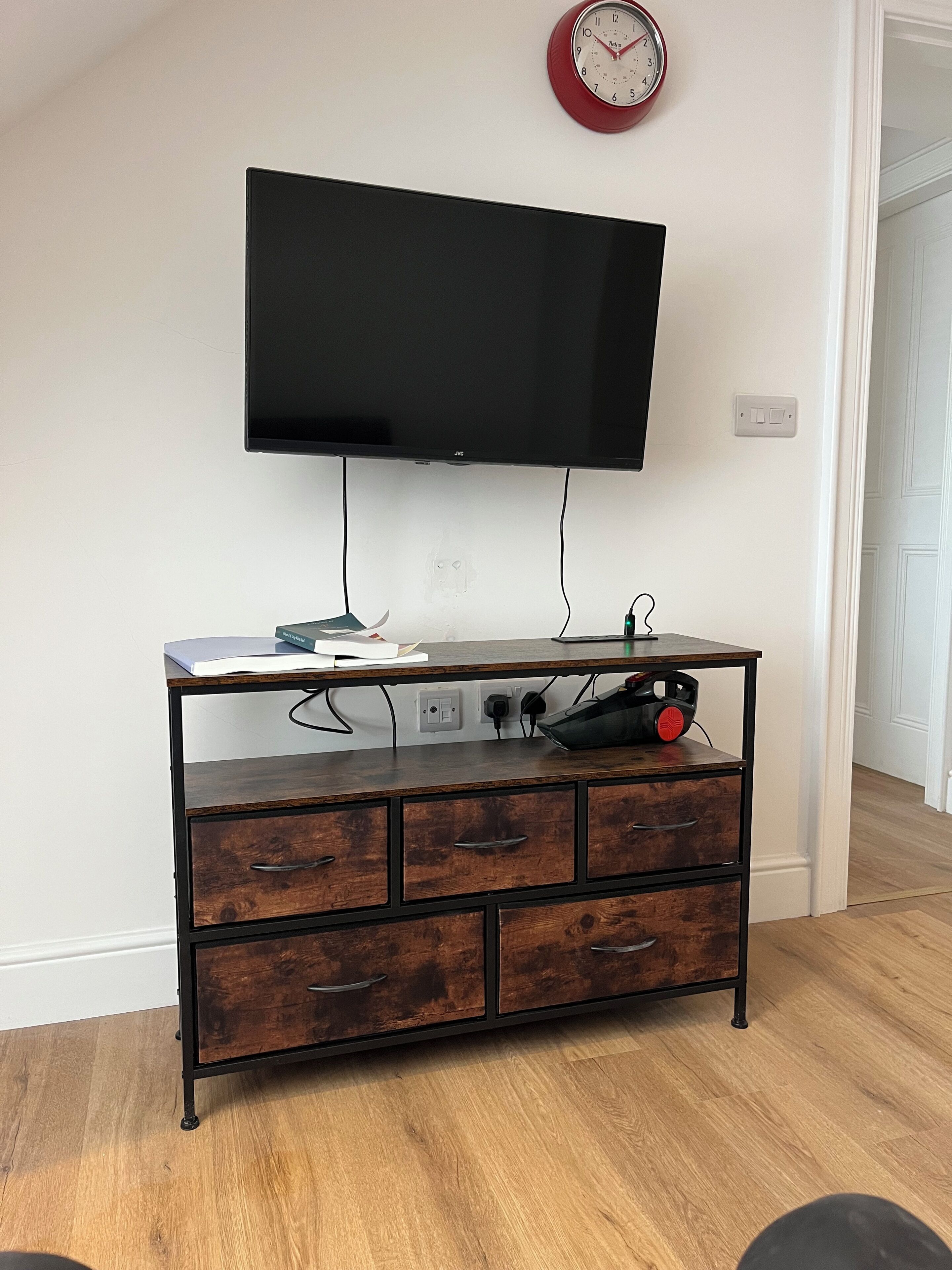 Una habitación con una televisión de pantalla plana montada en la pared, una mesa consola de madera con cajones y un reloj rojo en la pared.