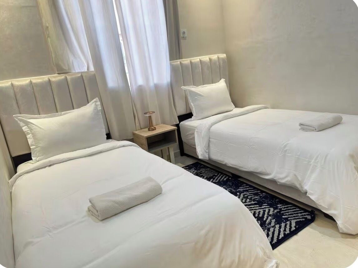 Une chambre d’hôtel avec deux lits, chacun doté de draps et d’oreillers blancs, une table de chevet avec une lampe et un tapis à motifs sombres.