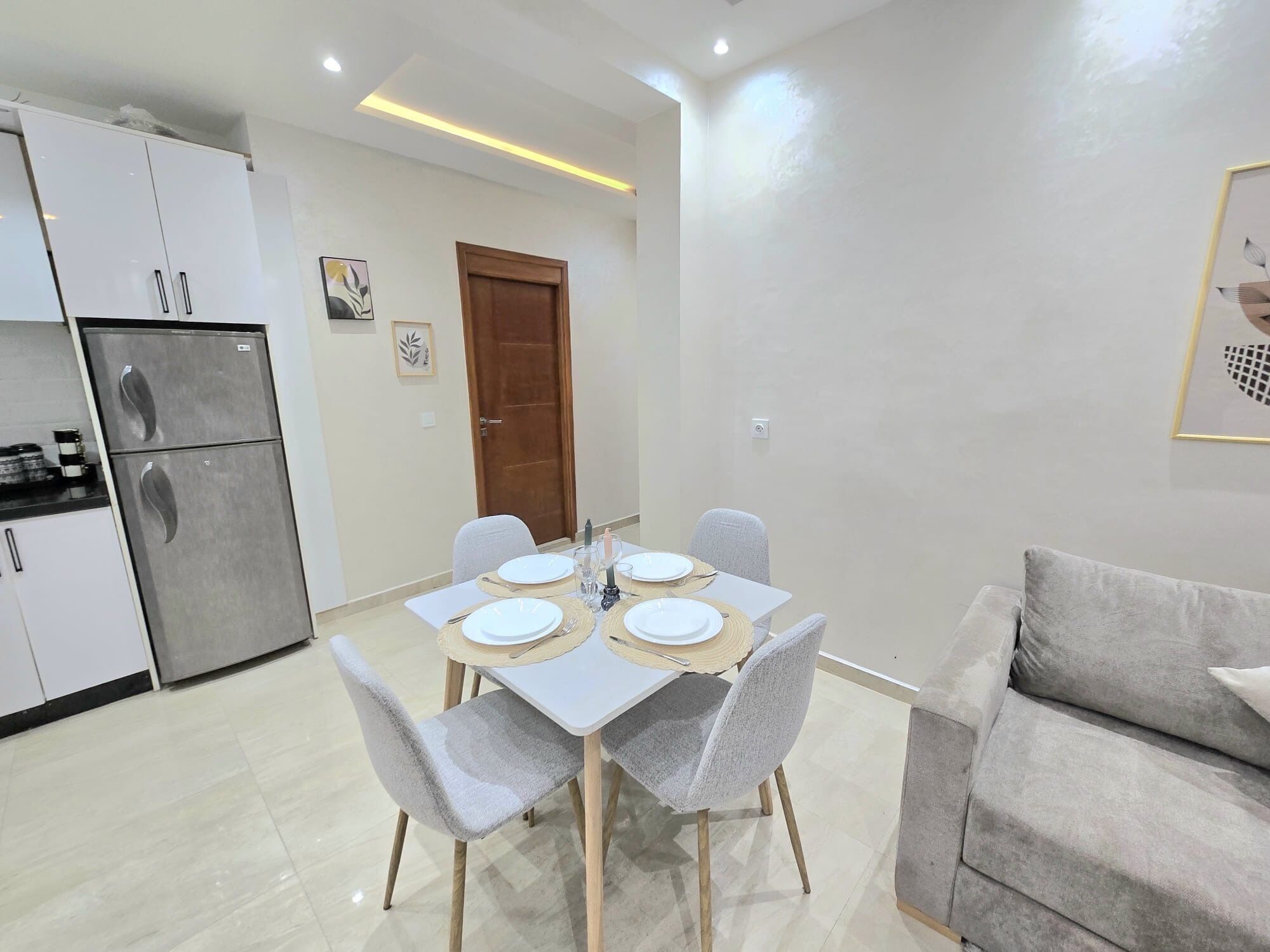A modern kitchen with a built-in refrigerator, a dining table set for four with placemats, and a grey sofa in the living area.