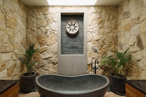 A stone-walled bathroom with a freestanding bathtub, a decorative wall panel, and two potted plants. - Villa Solaia by Alfred in Bali (Pecatu)