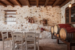 Una habitació de parets de pedra amb bigues de fusta, taula i cadires, tones de vi i estanteries plenes de botelles.