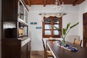 A dining area with a wooden table, chairs, a chandelier, and a window with curtains. - Villa Lithos by Your Villa (Aptera)