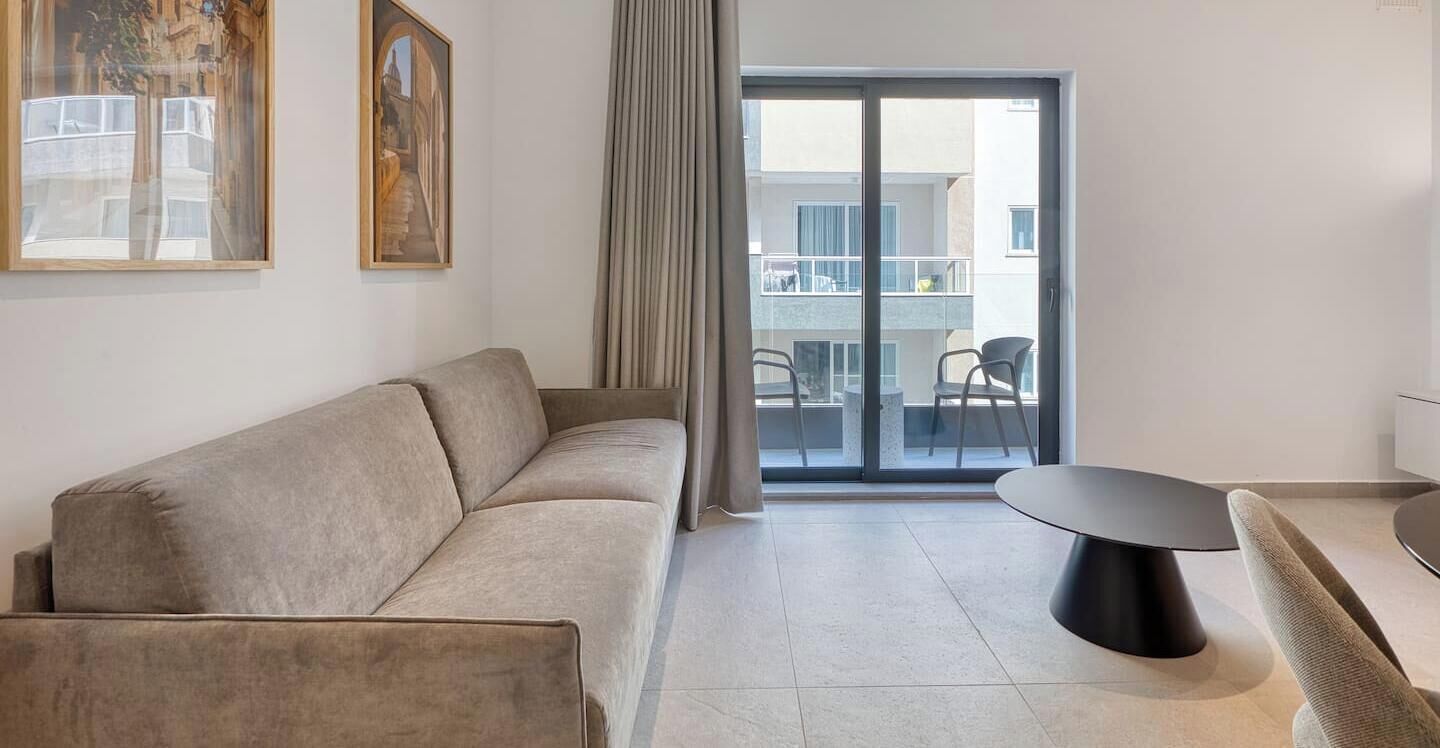 A modern living room with a sofa, a dining table, and a view through a glass door to an outdoor balcony.