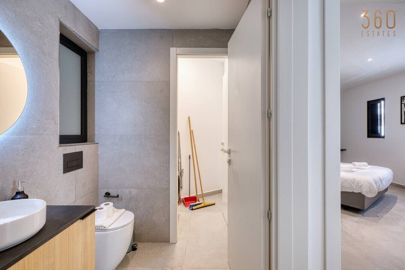 Un bagno moderno con uno specchio grande, un water e un lavandino. Una porta conduce a una camera da letto con un letto e un comodino.