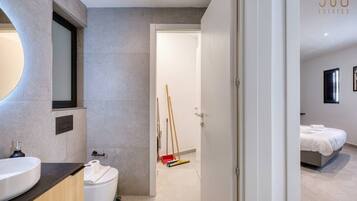 Un bagno moderno con uno specchio grande, un water e un lavandino. Una porta conduce a una camera da letto con un letto e un comodino.