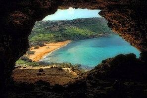 A coastal landscape viewed through a cave opening, featuring a beach, clear blue waters, and distant buildings. - Gozo Countryside Views | 3 BR Apt. Sannat w/AC (Sannat)