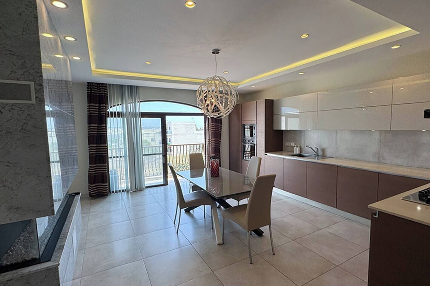 A modern kitchen with a dining area, a large window, and a decorative light fixture.