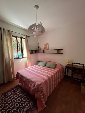 A bedroom with a bed, a window with curtains, a wall-mounted shelf with a guitar and picture, a bedside table with a lamp, and a patterned rug. - Looking for peace and tranquility in the heart of Balagne, Corsica? (Monticello)
