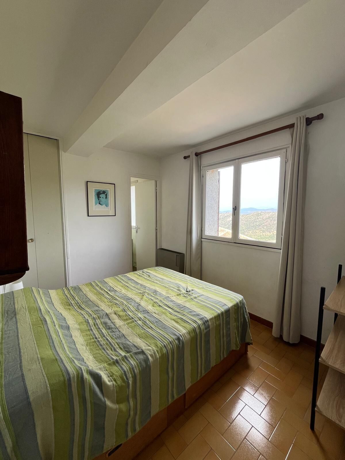 A bedroom with a bed, a window with curtains, a door, and a framed picture on the wall.