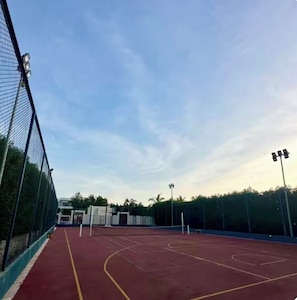 An outdoor basketball court with a red surface, surrounded by a chain-link fence, and equipped with floodlights. - Hawana Lagoons (ولاية طاقة،)
