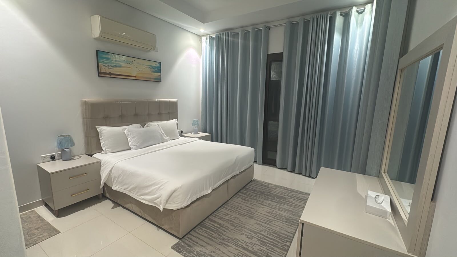 A modern hotel bedroom with a bed, nightstands, a bedside table, a mirror, and a window with curtains.