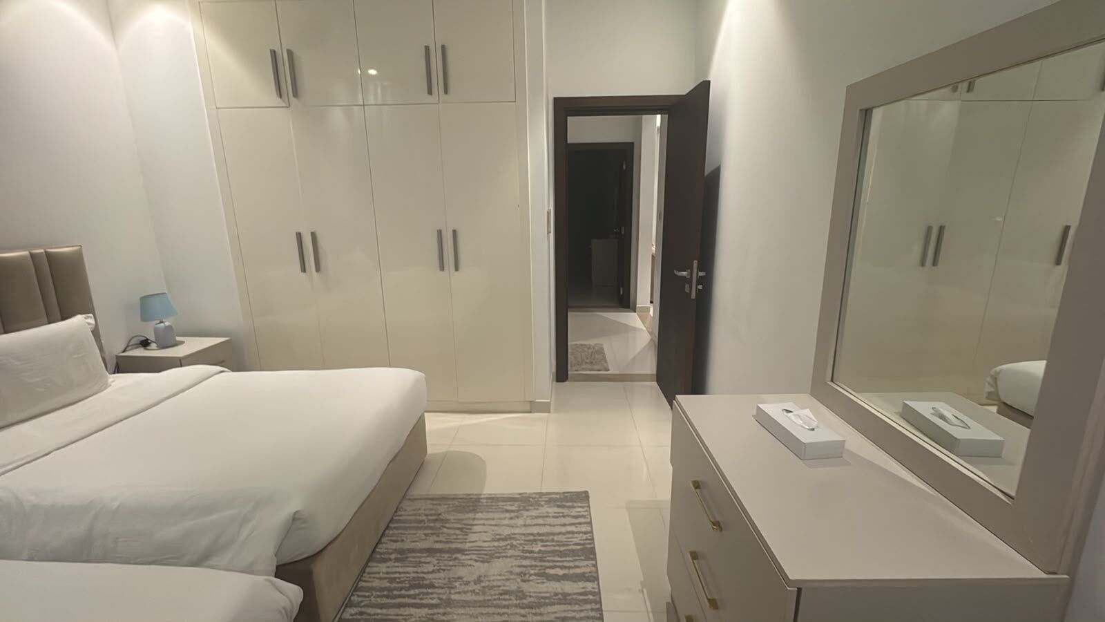 A modern bedroom with a large bed, bedside tables, a dresser, a mirror, and a walk-in closet.