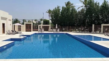 A rectangular swimming pool with a tiled edge, surrounded by a tiled deck. There are lounge chairs and umbrellas along the poolside.