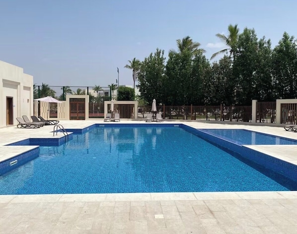 A rectangular swimming pool with a tiled edge, surrounded by a tiled deck. There are lounge chairs and umbrellas along the poolside. - Hawana Lagoons (ولاية طاقة،)