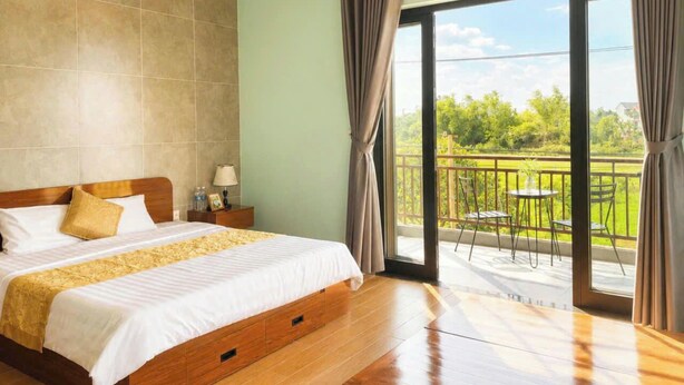 Ein Hotelzimmer mit einem großen Bett, einem Nachttisch, einer Lampe und Blick auf einen Balkon mit Außenmöbeln.