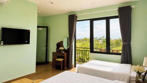 Ein Hotelzimmer mit zwei Betten, einem Schreibtisch mit Stuhl, einem Fernseher und einem Balkon mit Vorhängen.