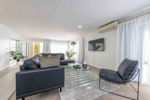 Ein modernes Wohnzimmer mit einer schwarzen Couch, einem Glastisch und einem schwarzen Sessel. An der Wand ist ein Fernseher angebracht, und das Zimmer verfügt über ein großes Fenster mit Vorhängen.