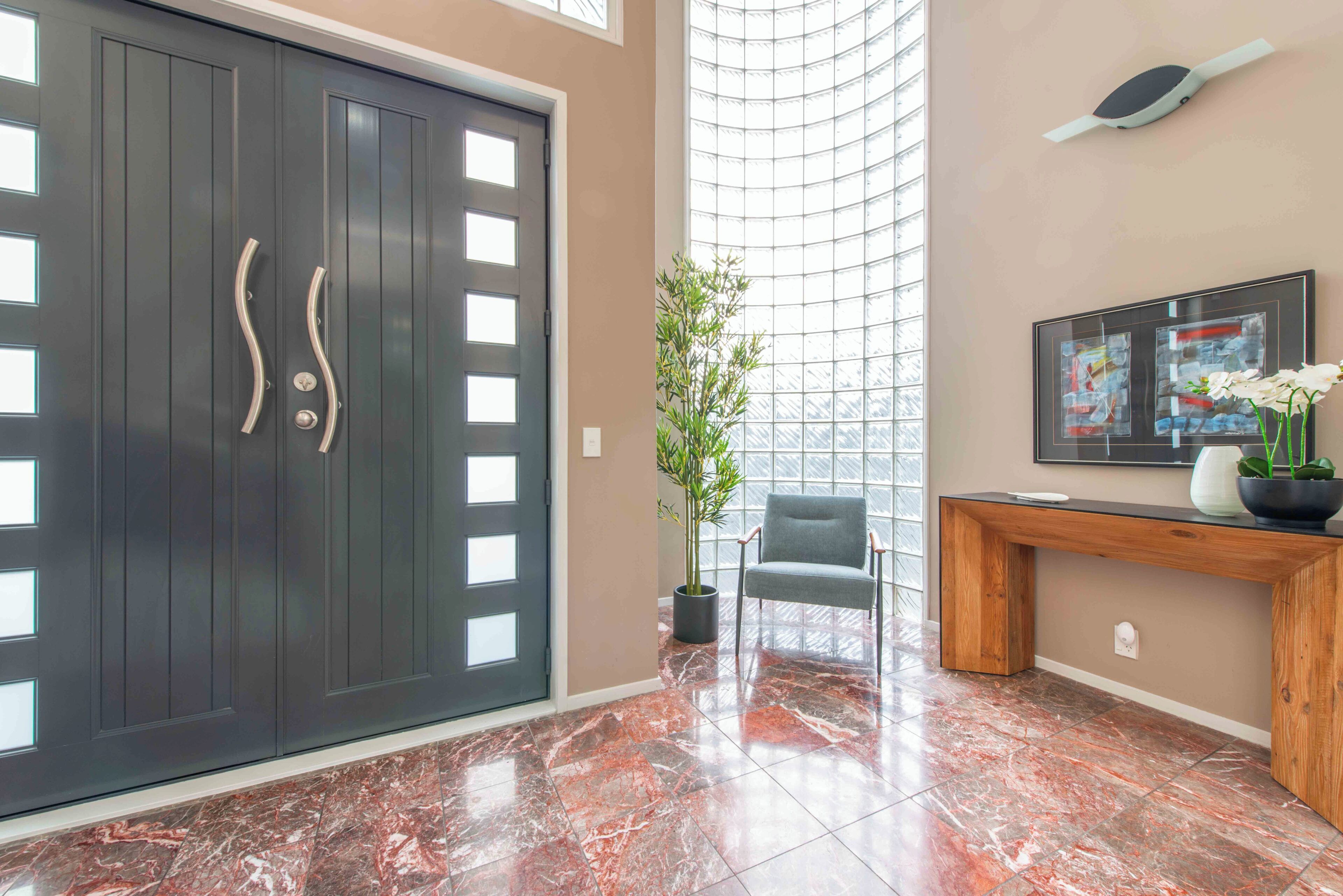 A modern interior with a dark wooden door, a potted plant, a grey chair, a wooden console table, and a framed abstract painting.